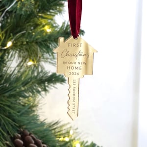 May include: A gold-colored, house-shaped key ornament hanging from a dark red velvet ribbon. The surface is engraved with the text FIRST Christmas IN OUR NEW HOME 2026 and 123 RANDOM STRET. The ornament has a reflective, metallic finish.