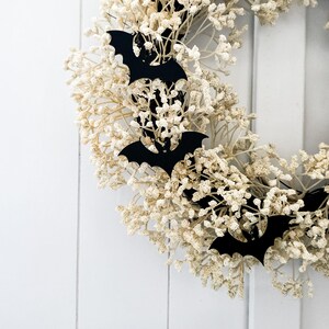 May include: A circular wreath composed of dried, cream-coloured flowers, decorated with black bat cutouts. The wreath is set against a white wooden background, creating a contrast between the natural elements and the Halloween theme.