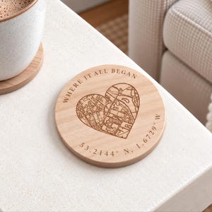 May include: A round light-colored wooden coaster featuring a laser-engraved heart shape containing a detailed street map. The text "WHERE IT ALL BEGAN" is arched above the heart, and the coordinates "53.2144° N, 1.6729° W" are printed below.