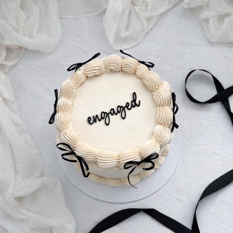 Pode incluir: Um bolo redondo creme decorado com glac&ecirc; e la&ccedil;os pretos. A palavra "engaged" est&aacute; escrita em preto na superf&iacute;cie do bolo. O bolo est&aacute; sobre uma superf&iacute;cie branca, com tecido branco e fita preta no fundo.