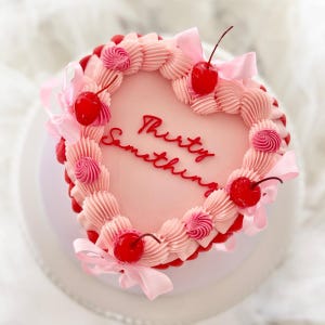 May include: A heart-shaped cake with pink icing and the words "Thirty Something" in red script. The cake is decorated with pink frosting swirls, red cherries, and pink ribbons. The cake sits on a white plate.