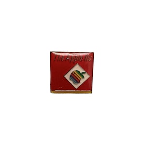 May include: A square metal enamel pin featuring a red background with the text The Apple llc in gold lettering. Centered is a white diamond shape containing a rainbow-striped apple graphic with a green leaf, finished with a gold border.