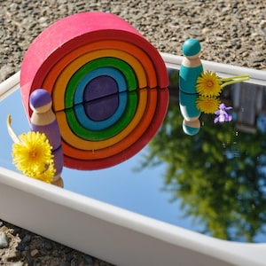 Può includere: Un impilatore arcobaleno in legno nei toni del rosso, arancione, giallo, verde, blu e viola. Due bambole di legno, una viola e una verde acqua, sono posizionate vicino all'impilatore. Sono presenti anche denti di leone gialli e un piccolo fiore viola.