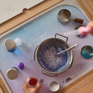 Puede incluir: Un conjunto de manualidades con una olla de acero inoxidable llena de líquido azul brillante, una cuchara y pequeños recipientes. La superficie tiene el texto "LET THE magic BEGIN". Varias herramientas e ingredientes están dispuestos para un proyecto creativo.