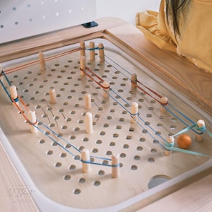 May include: A wooden board with a grid of holes and wooden pegs.  Elastic bands are stretched between the pegs, creating a maze-like pattern.  A small orange ball is in the maze.