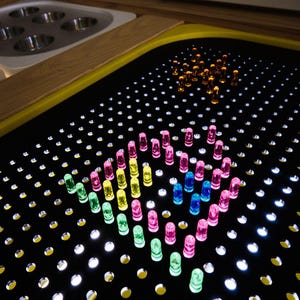 May include: A black light board with numerous small white lights and colorful translucent pegs arranged in a heart shape. Additional pegs are grouped in the upper right corner. A wooden and yellow border surrounds the board.