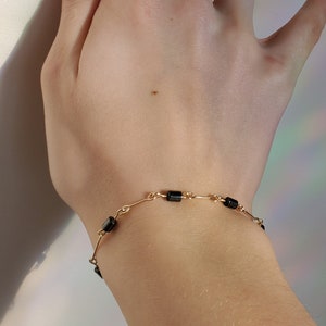 May include: A gold bracelet with black square beads. The bracelet is on a person's wrist.