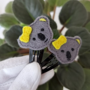 May include: Two gray felt koala hair clips with yellow felt bows. The clips are attached to black metal hair clips.