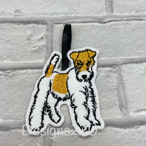 May include: White felt ornament of a wirehaired fox terrier dog with black embroidered details and a tan patch on its chest. The ornament has a black ribbon loop for hanging.