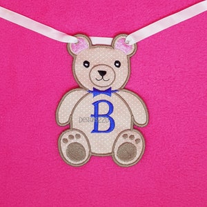 May include: A brown teddy bear with pink ears and a blue bow tie. The bear is sitting and has a blue letter "B" on its chest. The bear is on a pink background.