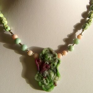 Könnte beinhalten: Ein grüner und rosafarbener geschnitzter Steinanhänger hängt an einer Halskette aus grünen, rosafarbenen und silbernen Perlen.