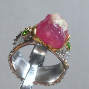 Może przedstawiać: Pierścionek z dużym różowym kamieniem szlachetnym osadzonym w złocie, otoczonym mniejszymi zielonymi kamieniami szlachetnymi i srebrnymi akcentami.