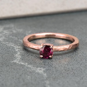May include: A hammered rose gold band ring with a deep red gemstone.