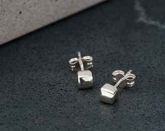 May include: A pair of silver stud earrings with a square shape. The earrings are on a dark gray surface.