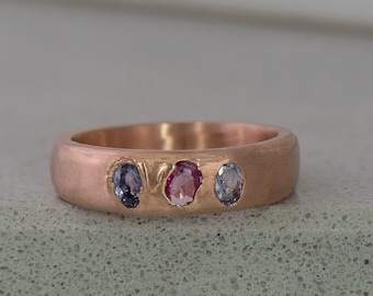 May include: A rose gold band ring with three oval-shaped gemstones. The gemstones are a light pink, a dark blue, and a light blue.