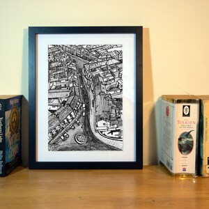 May include: A framed black and white print of a city street scene, flanked by books and a candle. The print is centered, with a detailed pen and ink drawing of buildings and a road. Books by Philip Pullman and J.R.R. Tolkien are visible.
