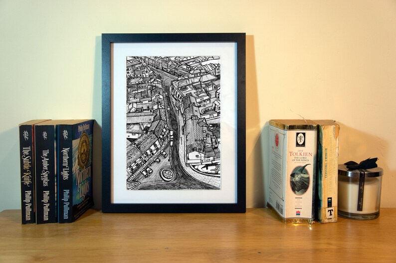 May include: A framed black and white print of a city street scene, flanked by books and a candle. The print is centered, with a detailed pen and ink drawing of buildings and a road. Books by Philip Pullman and J.R.R. Tolkien are visible.