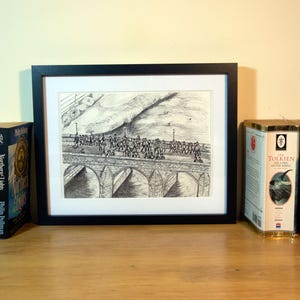 May include: A framed black and white print of a bridge scene, flanked by books and a candle. The books include titles like "The Subtle Knife" and "Northern Lights." A candle in a glass jar with a black bow sits nearby. The items are arranged on a wooden surface.