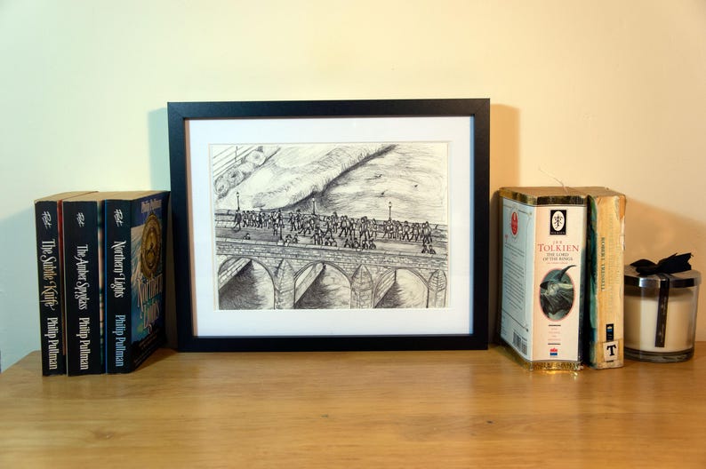 May include: A framed black and white print of a bridge scene, flanked by books and a candle. The books include titles like "The Subtle Knife" and "Northern Lights." A candle in a glass jar with a black bow sits nearby. The items are arranged on a wooden surface.
