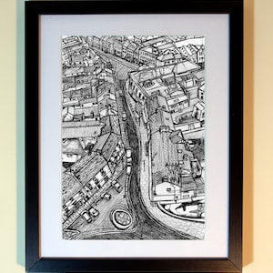 May include: Black and white pen and ink drawing of a city street scene, framed in a black frame with a white mat. The artwork depicts buildings, roads, and a roundabout from an aerial perspective.
