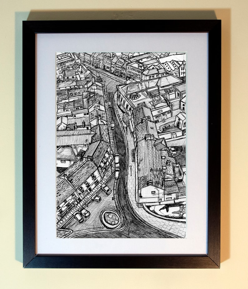 May include: Black and white pen and ink drawing of a city street scene, framed in a black frame with a white mat. The artwork depicts buildings, roads, and a roundabout from an aerial perspective.