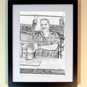 May include: A framed pencil sketch of a man seated at a bar, holding a pint of beer. The man wears a plaid shirt and is seated at a wooden table. The sketch is in a black frame with a white mat.
