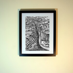May include: Black and white pen and ink drawing of a city street scene, framed in a black rectangular frame. The artwork depicts buildings, roads, and a roundabout, creating a detailed aerial view.