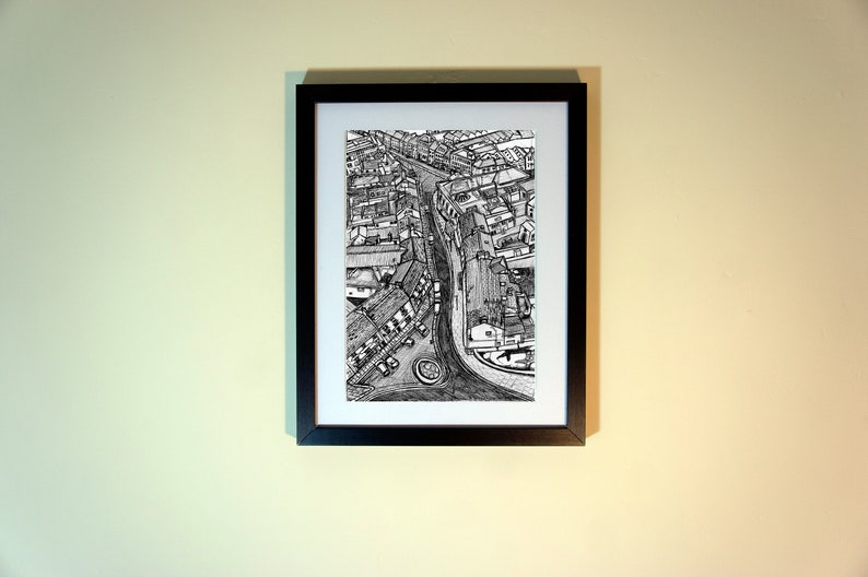 May include: Black and white pen and ink drawing of a city street scene, framed in a black rectangular frame. The artwork depicts buildings, roads, and a roundabout, creating a detailed aerial view.