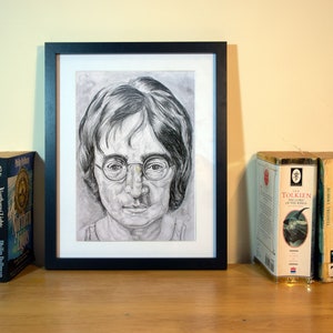 Pu&ograve; includere: Un ritratto a matita incorniciato di un uomo con occhiali rotondi. L'opera d'arte &egrave; esposta su una superficie di legno, accanto a libri di Philip Pullman e a "Il Signore degli Anelli" di J.R.R. Tolkien. &Egrave; presente anche una candela in un barattolo di vetro.