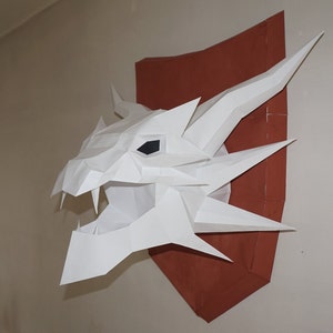May include: White paper dragon head sculpture mounted on a brown shield-shaped backing. The dragon has a fierce expression with sharp teeth and a single black eye.