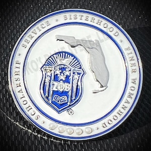 May include: A silver coin with a blue and white design. The coin features a crest with the letters "ZOB" and the year "1929" in the center. The coin also features a map of Florida and the words "Scholarship, Service, Sisterhood, Finer Womanhood".