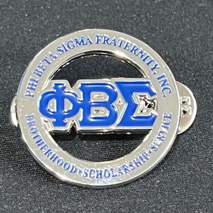 May include: A silver-coloured pin with a blue and silver emblem. The emblem features the Greek letters ΦΒΣ in blue, surrounded by a circular border with the text "PHI BETA SIGMA FRATERNITY, INC." and "BROTHERHOOD SCHOLARSHIP SERVICE."