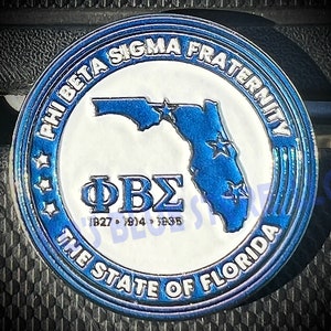 May include: A blue and white circular pin with the text "Phi Beta Sigma Fraternity" and "The State of Florida" around the edge. The center of the pin features a blue outline of the state of Florida with stars and the letters "ΦΒΣ" with the dates "1927, 1914, 1835" underneath.