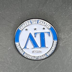 May include: A silver and blue Lambda Tau Chapter pin featuring the letters "AT" in blue on a white background. The pin also has three stars and the text "Florida International University #Elite".