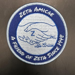 May include: A round, embroidered patch with a blue border and a light blue center. The patch features the words "ZETA AMICAE" and "A FRIEND OF ZETA SINCE 1948" in white text. A graphic of two hands shaking is in the center.