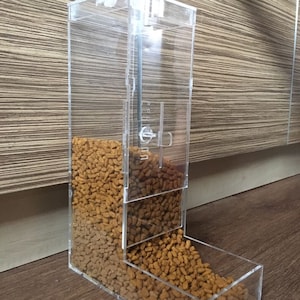 Puede incluir: Dispensador transparente de comida para mascotas de acrílico, lleno de croquetas secas. El dispensador tiene un diseño rectangular con una bandeja de alimentación en la parte inferior. Una varilla metálica es visible en el interior.