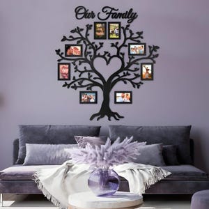 Pode incluir: Decoração de parede de árvore genealógica de madeira preta com oito molduras para fotos. O design da árvore inclui um tronco em forma de coração e galhos com as palavras "Our Family" no topo. A parede é pintada de roxo claro.