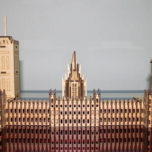 Può includere: Un modello in legno di un complesso di edifici, con una torre alta, una struttura centrale con una guglia e una torre dell'orologio. Il modello è realizzato in legno chiaro e presenta dettagli intagliati.