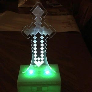 Pode incluir: Uma lâmpada em forma de espada pixelizada e brilhante. A espada de acrílico transparente assenta sobre uma base quadrada verde com duas luzes verdes. O design da espada é uma estética popular de videojogos, e a lâmpada emite uma luz ambiente suave.