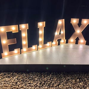 Op de afbeelding: Grote, verlichte marquee letters met de tekst "BELLA XV". De letters hebben een warme, lichtbruine kleur en zijn verlicht met kleine, ronde lampen. De display staat tegen een donkere achtergrond, wat duidt op een evenement of viering.