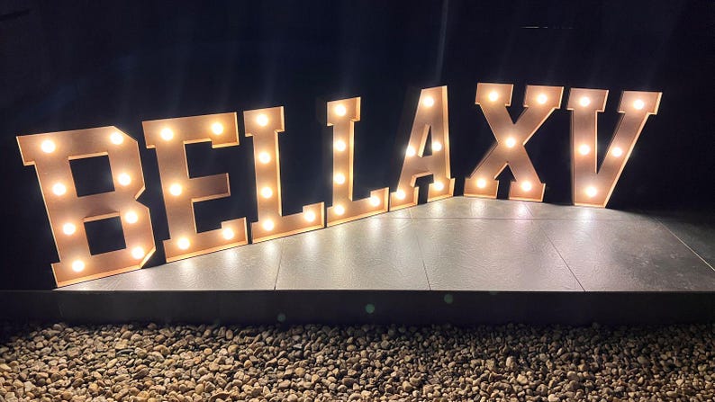 May include: Lit-up marquee letters spelling out "BELLA XV" in a warm, inviting glow. The letters are made of a light-colored material and are illuminated with small, round lights. The XV likely represents a milestone.