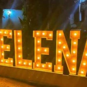 May include: Large illuminated sign spelling out the name "HELENA". The letters are a warm orange color and are lit with multiple small, round lights. The sign is likely for a special event or business.
