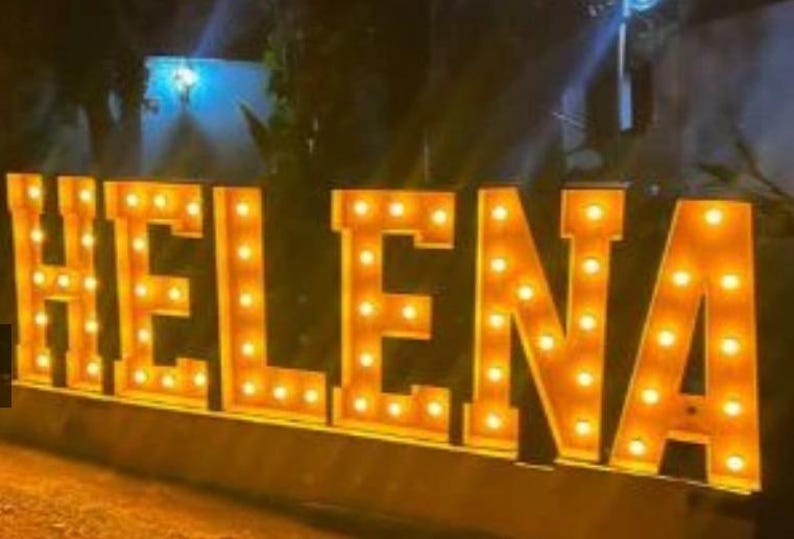May include: Large illuminated sign spelling out the name "HELENA". The letters are a warm orange color and are lit with multiple small, round lights. The sign is likely for a special event or business.