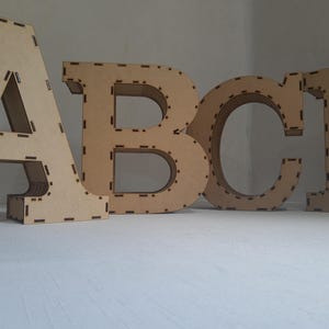 May include: Four large wooden letters, A, B, C, and I, stand upright. The letters are made of light brown wood with a cutout design. The letters are on a white surface, and the background is a light gray.