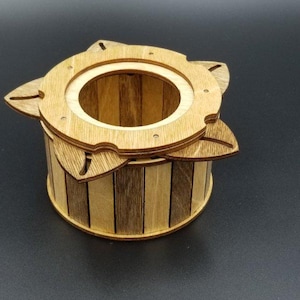 Peut inclure: Une boîte cylindrique en bois avec un couvercle. La boîte est faite de bandes de bois claires et foncées. Le couvercle a une ouverture circulaire et des accents en forme de feuille. La boîte est probablement destinée au stockage ou à la décoration.
