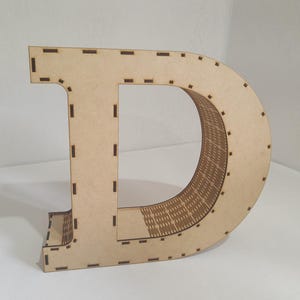 May include: A large, freestanding wooden letter "D" in a light brown color. The letter is constructed from multiple layers, with a detailed interior design. The edges have small, evenly spaced cutouts. The letter is likely used for home decor or crafting.