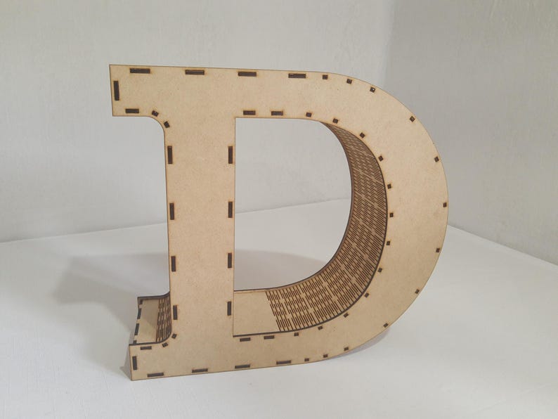 May include: A large, freestanding wooden letter "D" in a light brown color. The letter is constructed from multiple layers, with a detailed interior design. The edges have small, evenly spaced cutouts. The letter is likely used for home decor or crafting.