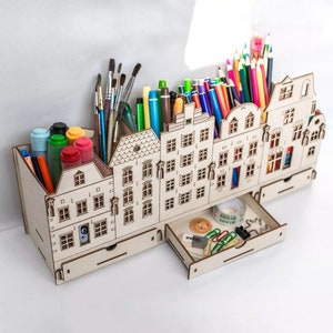May include: A wooden desk tidy in the shape of a street of houses. It contains compartments filled with art supplies such as paintbrushes, pens, and pencils. A small drawer holds stationery items like paperclips and tape.
