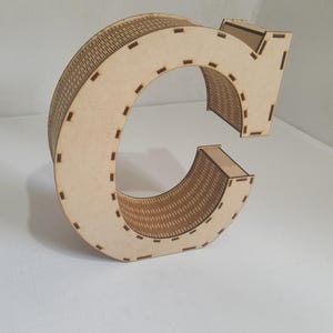May include: A large, wooden letter "C" with a decorative, woven-like pattern on the curved surfaces. The letter is made of light-colored wood and has a three-dimensional design. The edges are marked with small, evenly spaced cutouts.