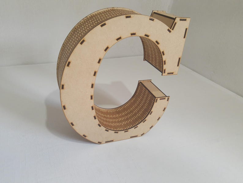 May include: A large, wooden letter "C" with a decorative, woven-like pattern on the curved surfaces. The letter is made of light-colored wood and has a three-dimensional design. The edges are marked with small, evenly spaced cutouts.